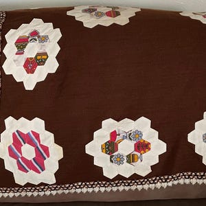 May include: A brown quilt with white, hexagon-shaped patches featuring colorful floral and geometric designs. The quilt has a crocheted border in white and brown, adding a handcrafted touch.