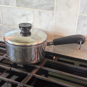 May include: A stainless steel saucepan with a black handle and lid, sitting on a black stovetop. The pot has a shiny, reflective surface and a black knob on the lid. The handle has a loop for hanging. The background features a light-colored tile backsplash.