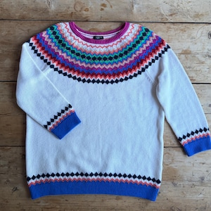 May include: A white sweater with a colorful geometric pattern across the chest and on the cuffs and hem. The pattern includes shades of red, blue, green, and pink. The sweater has a crew neck and long sleeves.