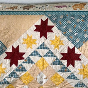May include: A handmade quilt with a patchwork design. The quilt features a central pattern of burgundy and yellow stars, set against a cream and blue plaid background. The border has animal and nature motifs.