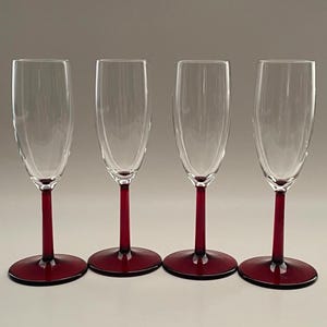 May include: Four clear champagne flutes with ruby red stems and bases. The glasses have a classic, elegant design, perfect for celebratory occasions. The stems and bases are a deep, rich red color, contrasting with the clear glass bowls.