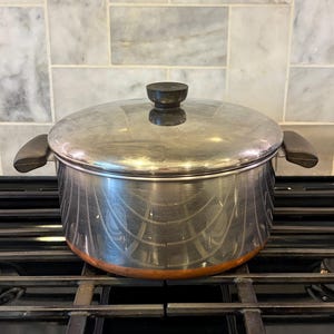 May include: A stainless steel stockpot with a matching lid, featuring two dark brown side handles and a dark brown knob on the lid. The pot has a polished silver exterior finish with a visible copper-colored base band, designed for stovetop use.
