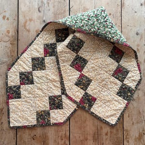 May include: A quilted table runner with a cream-colored background and a repeating pattern of dark squares with floral designs. The border and the underside feature a green and red holly pattern. The runner is placed on a wooden surface.
