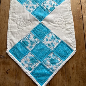 May include: A quilted table runner with a diamond pattern. The diamonds alternate between solid turquoise and floral print fabric. The runner has a white border and white quilted floral designs on the white sections.
