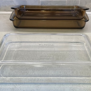 May include: Three rectangular glass baking dishes. Two are amber-colored and stacked, while the third is clear and sits in front. The dishes are made of a durable, heat-resistant glass, suitable for oven use. The clear dish has a lid.