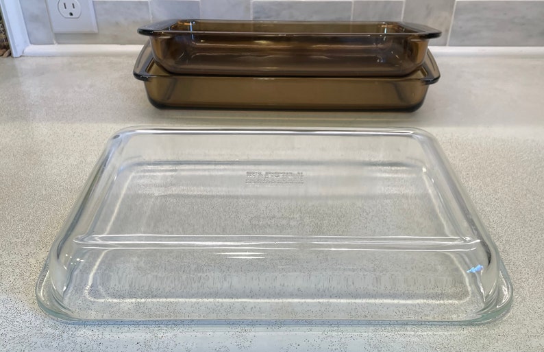 May include: Three rectangular glass baking dishes. Two are amber-colored and stacked, while the third is clear and sits in front. The dishes are made of a durable, heat-resistant glass, suitable for oven use. The clear dish has a lid.
