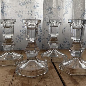 May include: Four clear glass candle holders with a faceted design. The candle holders have a hexagonal base and a flared top. The background includes a floral patterned curtain and a wooden surface.