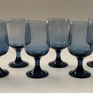May include: Set of six blue-tinted stemmed glasses. The glasses have a tulip-shaped bowl and a dark blue base and stem. The glasses are arranged in a row, showcasing their elegant design.