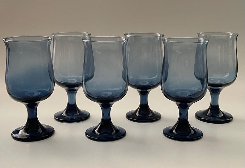 May include: Set of six blue-tinted stemmed glasses. The glasses have a tulip-shaped bowl and a dark blue base and stem. The glasses are arranged in a row, showcasing their elegant design.