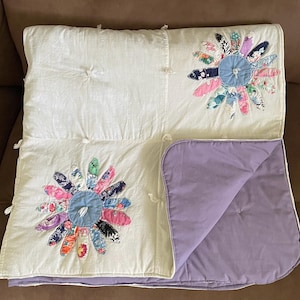 May include: A handmade quilt featuring two large floral appliqués with colorful petal designs on a white background. The quilt has a lavender backing and is tied with white yarn. The overall design is a patchwork style.