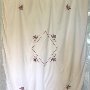 May include: A rectangular white fabric tablecloth featuring a central diamond-shaped outline embroidered in dark red thread. Small floral motifs in red and green are embroidered at the four corners and around the central diamond shape.