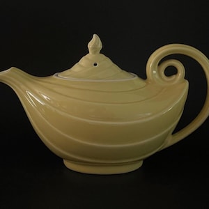 May include: A pale yellow ceramic teapot with a matching lid. The teapot has a curved handle and spout, and a decorative swirl pattern. The lid has a small finial. The teapot is set against a black background.