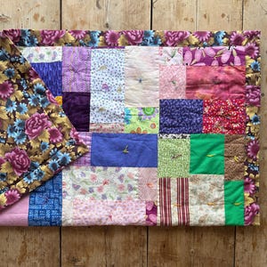 May include: A patchwork quilt with a floral border and a variety of colorful fabric squares. The quilt features shades of pink, purple, blue, and green. The border has a brown background with pink and purple flowers.