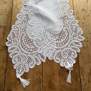 May include: White lace table runner with intricate floral and geometric patterns. The runner has a scalloped edge and two tassels at the ends. This decorative textile is suitable for a dining table or dresser.