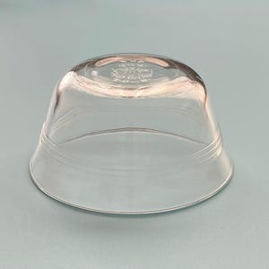 May include: A clear, inverted glass bowl with a wide base and a slightly curved top. The bowl has a decorative emblem etched on the top and two horizontal etched lines. The bowl is made of transparent glass.