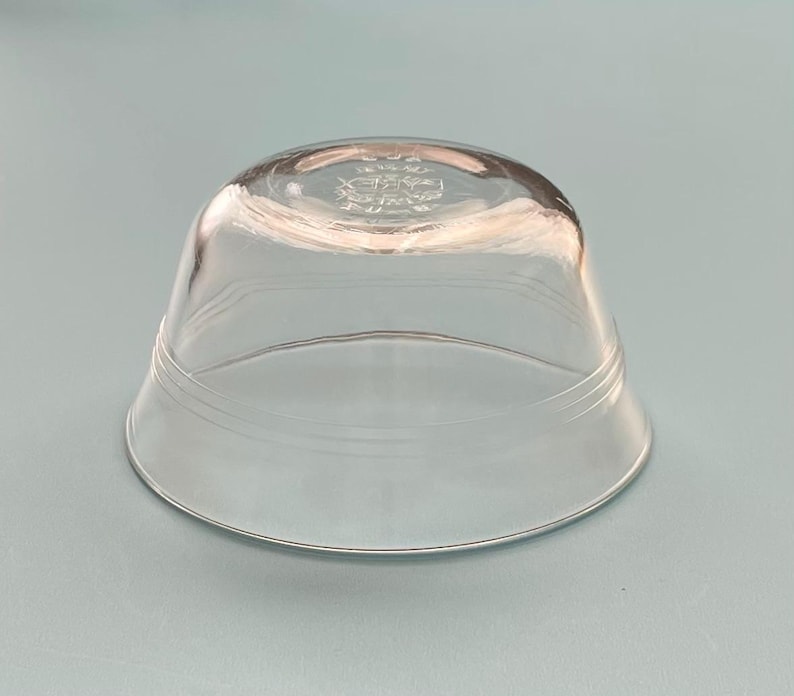 May include: A clear, inverted glass bowl with a wide base and a slightly curved top. The bowl has a decorative emblem etched on the top and two horizontal etched lines. The bowl is made of transparent glass.
