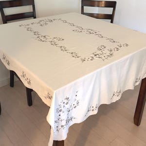 May include: A square white tablecloth featuring a dark gray embroidered floral pattern arranged in a rectangular border on the top surface, with additional matching floral embroidery along the scalloped edges of the fabric drape.