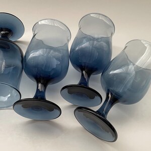 May include: A set of six blue-tinted stemmed glasses. The glasses have a rounded bowl shape and a dark blue base and stem. The glass color fades from a lighter blue at the top to a darker blue at the base.