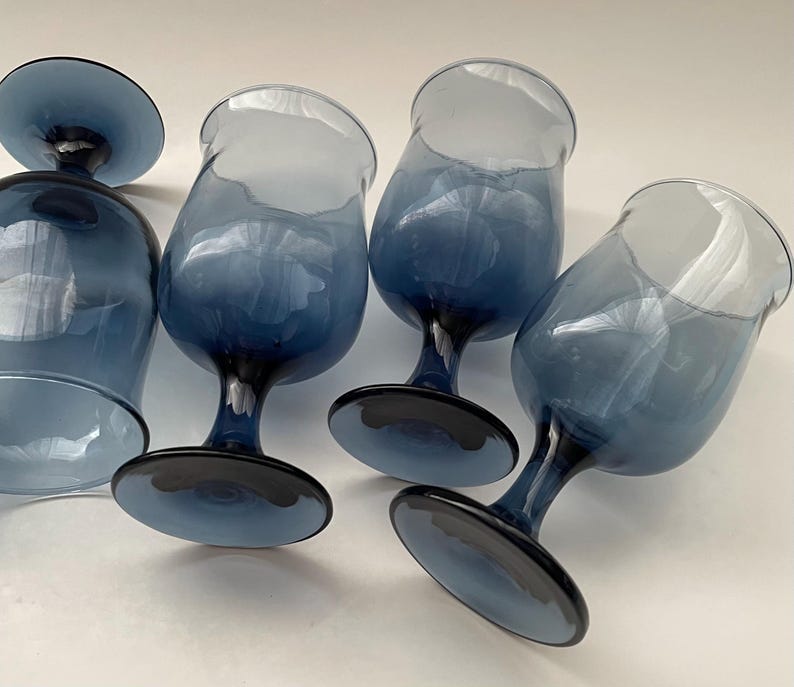May include: A set of six blue-tinted stemmed glasses. The glasses have a rounded bowl shape and a dark blue base and stem. The glass color fades from a lighter blue at the top to a darker blue at the base.