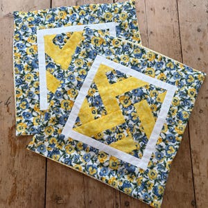 May include: Two square quilted pillows with a floral pattern of blue and yellow flowers on a white background. Each pillow has a central yellow geometric design framed by a white border.