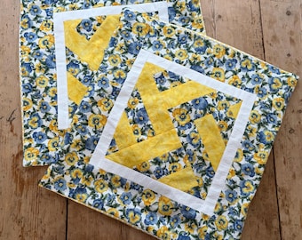 May include: Two square quilted pillows with a floral pattern of blue and yellow flowers on a white background. Each pillow has a central yellow geometric design framed by a white border.