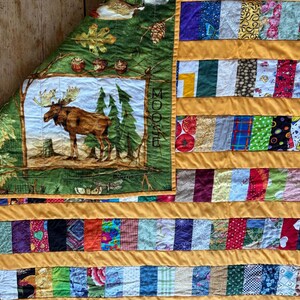 May include: A handmade quilt with a moose scene and a patchwork design. The moose is brown with large antlers, set against a forest backdrop. The quilt features a green border with the word "MOOSE" and colorful fabric strips.