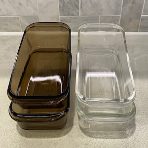 May include: Two sets of glass storage containers. One set is clear glass and the other is brown glass. Both sets are stacked on top of each other.