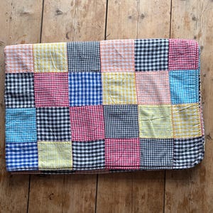 May include: A patchwork quilt with a variety of gingham squares in shades of pink, blue, yellow, black, and white. The quilt is made of fabric squares sewn together with yellow thread. The quilt is folded on a wooden surface.