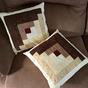 May include: Two square throw pillows with a patchwork design. The pillows feature a log cabin pattern in shades of brown, beige, and cream, with a small red square at the center. The pillows are on a brown sofa.