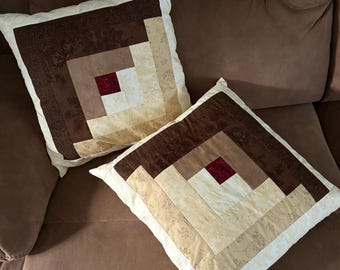 May include: Two square throw pillows with a patchwork design. The pillows feature a log cabin pattern in shades of brown, beige, and cream, with a small red square at the center. The pillows are on a brown sofa.