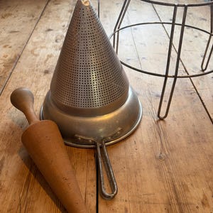 May include: A conical metal food mill with a fine mesh surface, a metal handle, and a matching metal wire stand. Beside the food mill is a smooth, light brown wooden pestle with a tapered, conical shape designed for pressing food through the mesh.