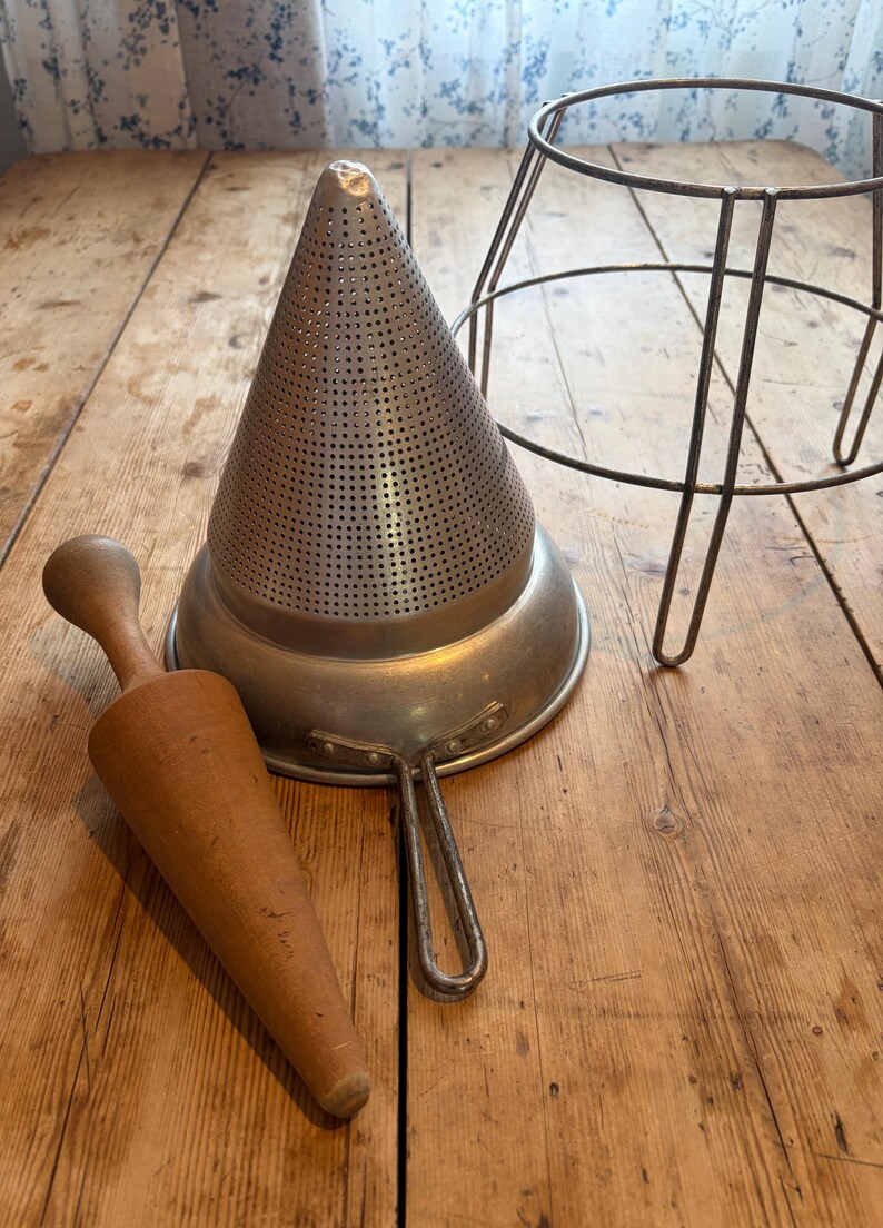 May include: A conical metal food mill with a fine mesh surface, a metal handle, and a matching metal wire stand. Beside the food mill is a smooth, light brown wooden pestle with a tapered, conical shape designed for pressing food through the mesh.