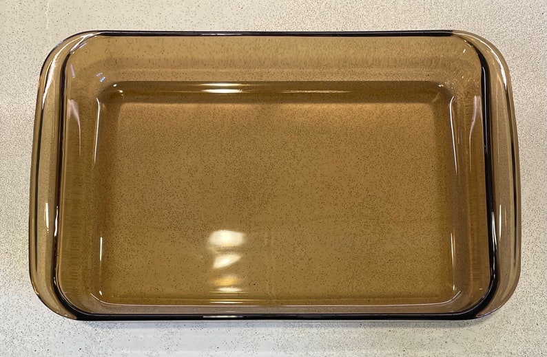 May include: Rectangular amber-colored glass baking dish with rounded corners and a slightly raised rim. The dish is empty and appears clean, suitable for baking or serving. The glass has a speckled appearance.