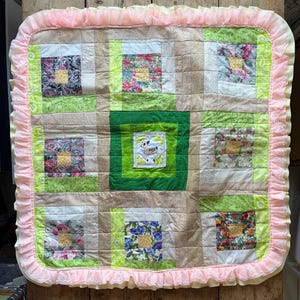 May include: A handmade quilt with a patchwork design. The quilt features squares of floral and patterned fabrics in shades of pink, green, and gold. A central square has an embroidered sheep. The quilt is trimmed with a pink ruffled border.