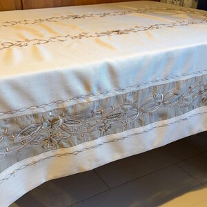 May include: A rectangular white tablecloth featuring a wide, horizontal band of light gray fabric along the edges. The fabric is decorated with intricate, swirling floral embroidery in metallic gold and silver thread.