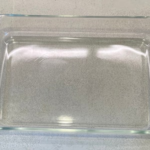 May include: A clear, rectangular glass baking dish with rounded corners. The dish is empty and sits on a speckled gray surface. Ideal for baking casseroles, desserts, and other dishes.