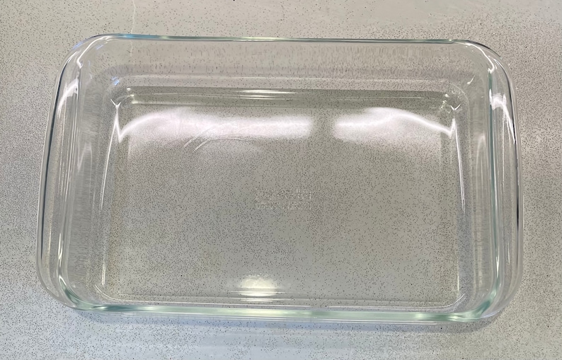 May include: A clear, rectangular glass baking dish with rounded corners. The dish is empty and sits on a speckled gray surface. Ideal for baking casseroles, desserts, and other dishes.