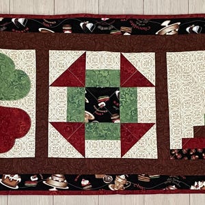 May include: A quilted table runner with three distinct sections. The first features a four-leaf clover design in red and green. The second has a geometric pattern with red and green triangles. The third section has a log cabin design in red and green.