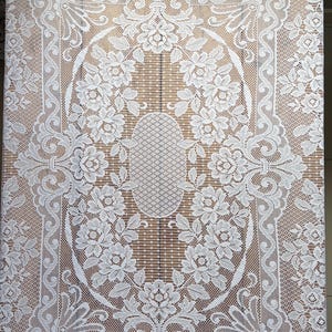 May include: White lace tablecloth with a floral design. The tablecloth features an oval floral centerpiece and a detailed border. The intricate lace pattern is visible throughout the design, creating a delicate and elegant look.