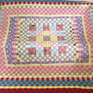 May include: A colorful patchwork quilt with a variety of patterns and colors. The quilt features squares of pink, purple, blue, yellow, and red. The quilt has a checkerboard design and is a handmade item.