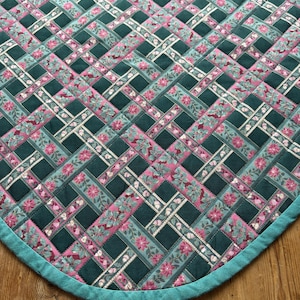 May include: A quilted textile with a woven pattern. The design features dark teal, pink floral, and light teal patterned strips. The edge is finished with a teal border. The textile is placed on a wooden surface.