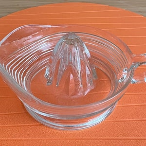 May include: A clear glass citrus juicer with a built-in reamer and a pouring spout. The juicer has a handle and is designed with a ribbed interior for efficient juice extraction. The juicer sits on an orange surface.