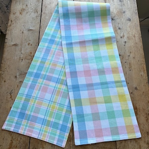 May include: A pastel-colored plaid table runner. The runner features a pattern of intersecting lines in shades of blue, pink, yellow, and green. The fabric is folded over on a wooden surface.