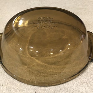 May include: A vintage amber-colored glass casserole dish with two side handles. The dish has a rounded shape and a smooth, reflective surface. The bottom of the dish has embossed text. A classic piece of kitchenware.
