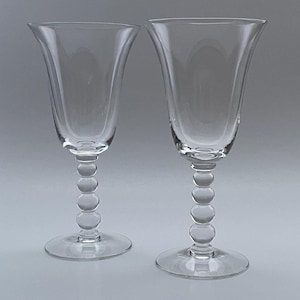 May include: Two clear, stemmed wine glasses. The bowls flare outward, and the stems feature a series of round, bubble-like accents. The glasses sit on a round base. The background is a solid, light gray.
