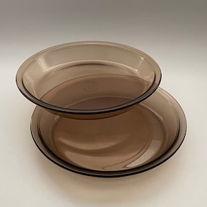 May include: Two brown glass pie plates stacked on top of each other. The top plate has a raised rim and the bottom plate has a flat rim.