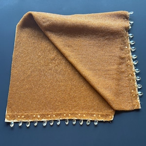 May include: A folded, golden-brown beaded textile with a decorative border. The border features small, clear, teardrop-shaped embellishments. The textile is likely a decorative item, such as a scarf or table runner.