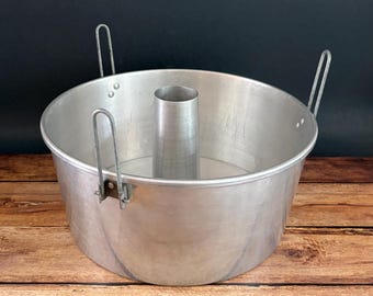 May include: A vintage aluminum angel food cake pan with a central tube and two handles. The pan is round with a fluted design and a light silver color. The handles are attached to the side of the pan.