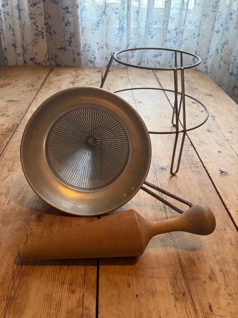 May include: A vintage food mill set consisting of a circular metal strainer with a fine mesh cone, a wire frame stand, and a tapered wooden pestle. The metal components have a matte finish, and the wooden pestle features a smooth, rounded handle.