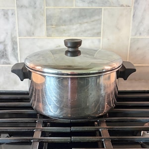 May include: A stainless steel cooking pot with a lid and black handles sits on a black stovetop. The pot is round with a shiny, reflective surface. The lid has a black knob. The background features a marble tile backsplash.
